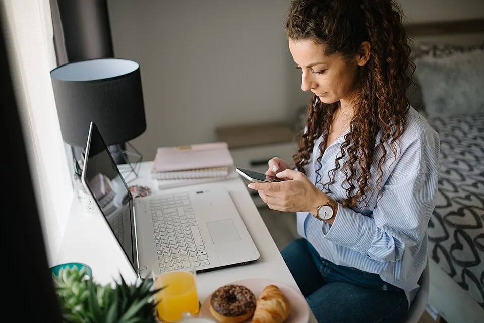 A woman checks a smartphone while working at a laptop at home.