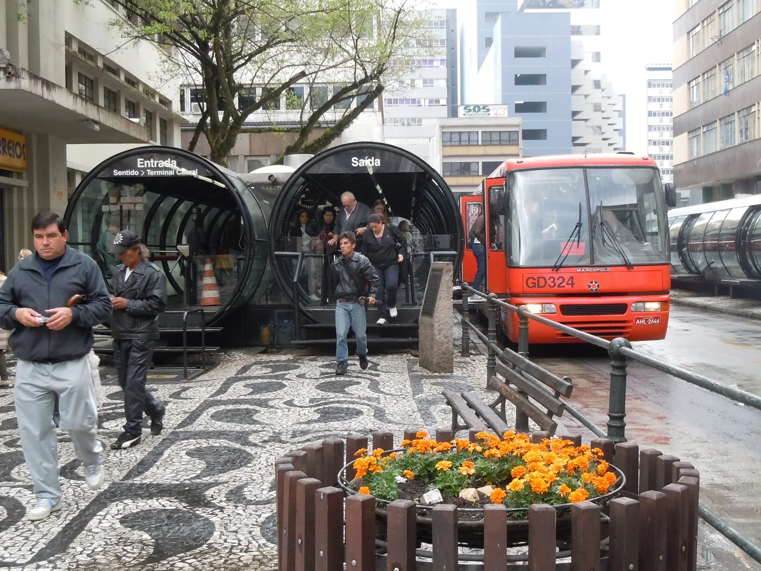 People leave a glass tube bus station beside a red bus in Curitiba, Brazil.