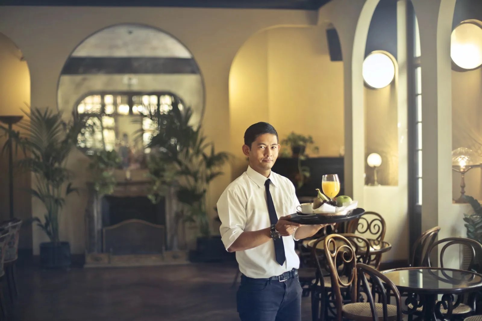 A waiter in a white shirt and dark apron carries a tray of drinks through a warmly lit restaurant.